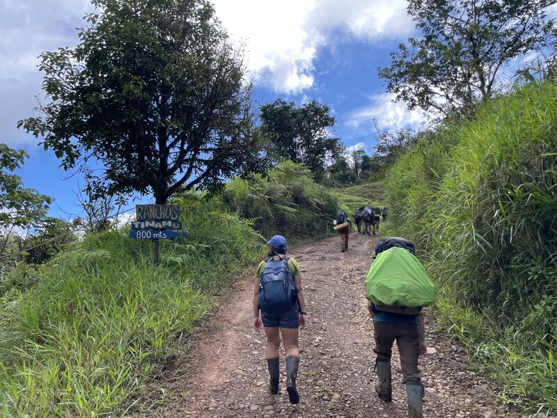 A group of hikers is walking up a dirt path on a grassy hillside. The hikers are wearing backpacks and hiking boots. There is a sign on the left side of the path. The sky is cloudy and the sun is shining through the clouds.